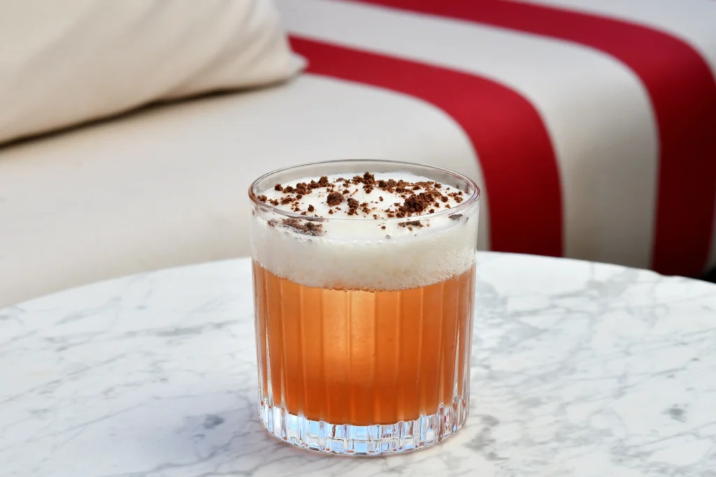 A glass of frothy iced coffee or cocktail topped with chocolate shavings sits on a white marble table with a striped sofa in the background.